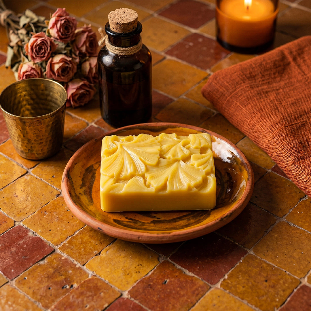 Bar of soap on a wooden plate with a bottle, cup, and candle on a tiled surface