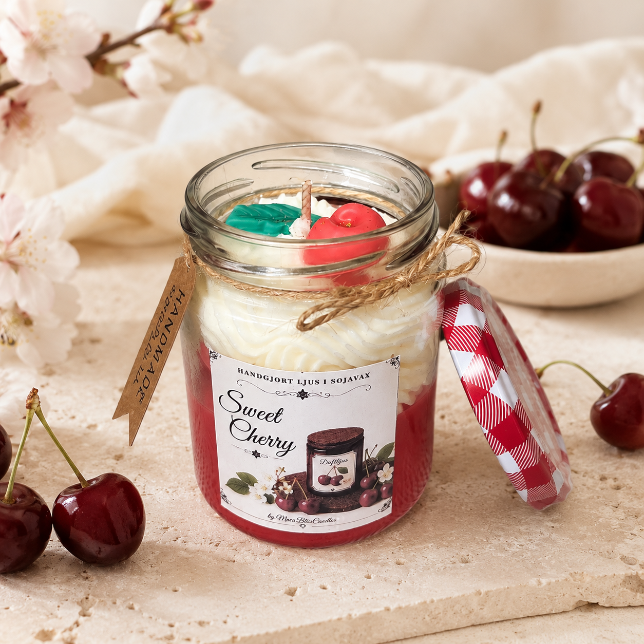 A jar candle labeled 'Sweet Cherry' with red and white layers, a green wax insert, and a red-and-white gingham lid leaning beside it on a stone surface. The scene is styled with cherries, soft cream fabric, and blurred blossoms in the background.